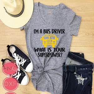 Puede incluir: Camiseta gris de cuello en V con el texto "Soy conductor de autobús, ¿cuál es tu superpoder?" y un gráfico de autobús escolar amarillo. La camiseta se muestra sobre una superficie de madera con un par de zapatillas negras, un sombrero de paja y unos vaqueros azules.