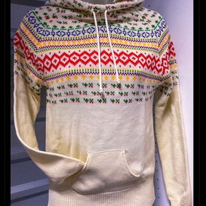 May include: A cream-colored hooded sweater with a colorful geometric pattern. The sweater has a front pocket and a drawstring hood.
