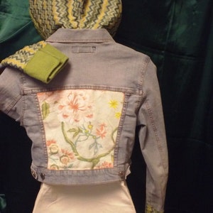 May include: A light blue denim jacket with a floral patterned fabric patch on the back. The jacket has a green and yellow chevron patterned hood.