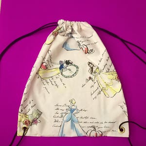 May include: White drawstring backpack featuring illustrations of Disney princesses in their iconic gowns. The bag has a repeating pattern of Cinderella, Belle, and Snow White, with script text. Black drawstrings and gold grommets complete the design.