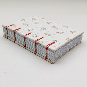 Op de afbeelding: Een klein, wit notitieboekje met een bloemenpatroon op de omslag. Het notitieboekje is gebonden met rode draad en heeft een witte rug.