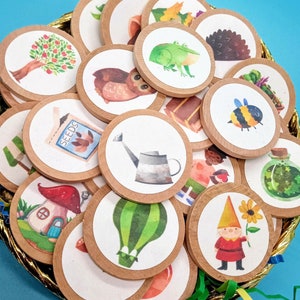May include: A set of 16 colorful round paper circles with various illustrations, including a frog, a gnome, a mushroom, a watering can, a bee, and a garden. The circles are arranged in a circular pattern on a blue background.
