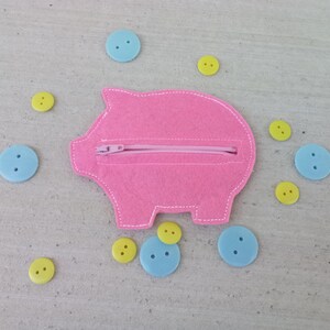 May include: A pink felt piggy bank with a zipper closure, surrounded by light blue and yellow buttons. The piggy bank is in the shape of a pig with white stitching around the edges.