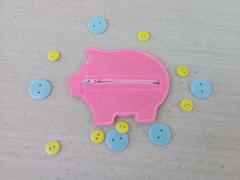 May include: A pink felt piggy bank with a zipper closure, surrounded by light blue and yellow buttons. The piggy bank is in the shape of a pig with white stitching around the edges.
