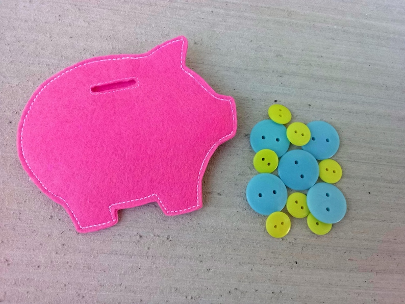 May include: A bright pink felt piggy bank with a slot for coins, next to a collection of blue and yellow buttons. The piggy bank is outlined with white stitching.