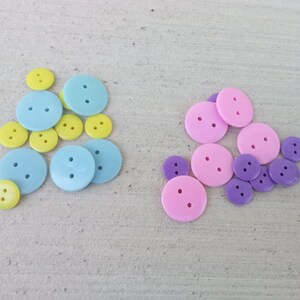 May include: An assortment of colorful buttons in various sizes. The buttons are in three color schemes: light blue and yellow, and pink and purple. Each button has two holes for sewing.