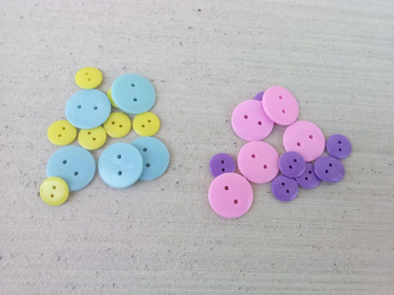 May include: An assortment of colorful buttons in various sizes. The buttons are in three color schemes: light blue and yellow, and pink and purple. Each button has two holes for sewing.