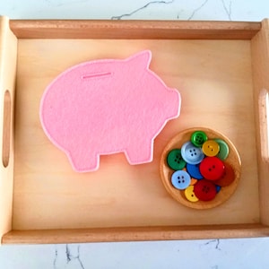 May include: A light pink felt piggy bank and a small wooden bowl filled with colorful buttons sit on a light wood tray. The buttons are various colors, including red, blue, green, and yellow. The tray has a rectangular shape with rounded corners.