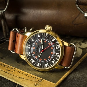 Puede incluir: Un reloj de pulsera dorado con una esfera negra y marcas blancas. El reloj tiene una correa de cuero marrón y una aguja de segundos roja. La esfera del reloj tiene la palabra "Aviator" impresa.