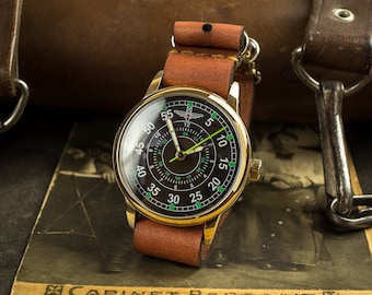 Könnte beinhalten: Eine goldfarbene Armbanduhr mit schwarzem Zifferblatt und grünen Markierungen. Die Uhr hat ein braunes Lederarmband und eine weiße Aufschrift mit der Aufschrift "CABINET-PORTRAIT".