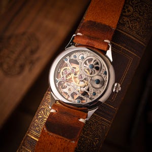 May include: A silver skeleton watch with a brown leather strap. The watch face displays intricate gears and mechanisms. The watch is set against a backdrop of a vintage book, showcasing a classic and detailed design.