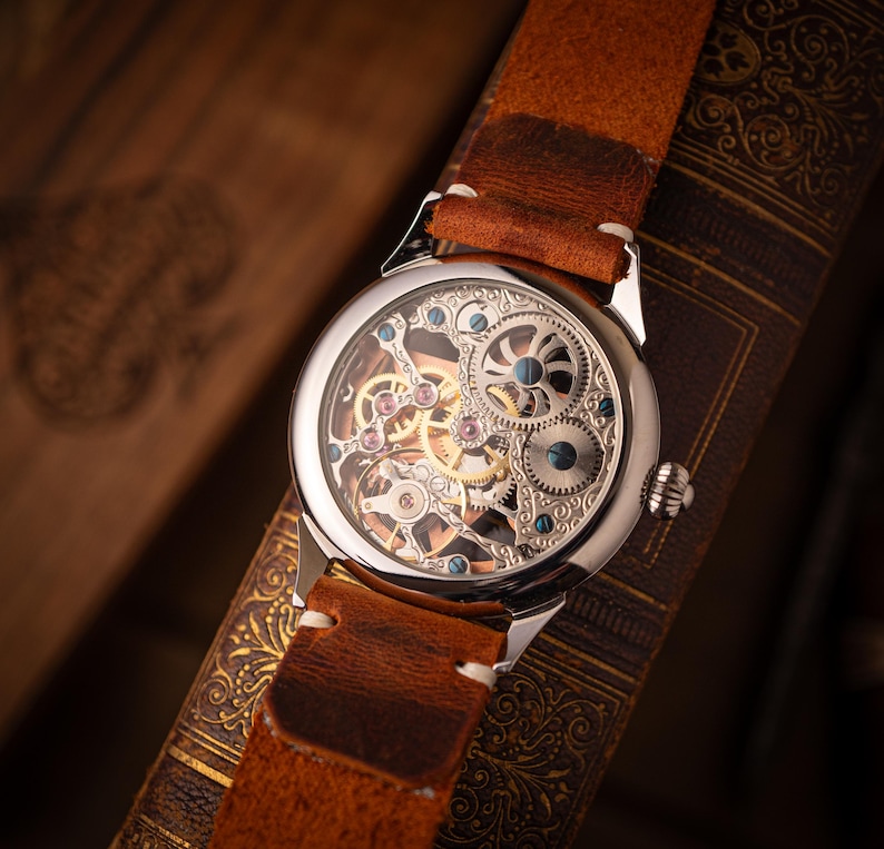 May include: A silver skeleton watch with a brown leather strap. The watch face displays intricate gears and mechanisms. The watch is set against a backdrop of a vintage book, showcasing a classic and detailed design.