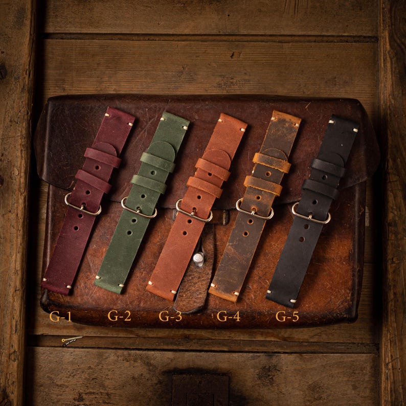 Puede incluir: Cinco correas de reloj de cuero en diferentes colores, incluyendo marr&oacute;n, verde y negro. Las correas est&aacute;n dispuestas sobre una superficie de cuero marr&oacute;n.