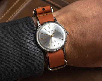 Könnte beinhalten: Eine silberne Armbanduhr mit weißem Zifferblatt und goldenen Akzenten. Die Uhr hat ein braunes Lederarmband und trägt die Aufschrift "Poljot de Luxe" und "23 jewels shockproof" auf dem Zifferblatt.