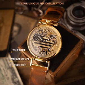 May include: Gold-toned watch with a brown leather strap. The watch face features a skeletonized design with an American flag motif and the text "THANK YOU FOR YOUR SERVICE". The watch includes the text "TOMMY DINSOU" and "2025". The watch is displayed on a wooden box.