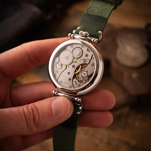 May include: A silver pocket watch with a green leather strap. The watch face is open, revealing the intricate inner workings of the watch.