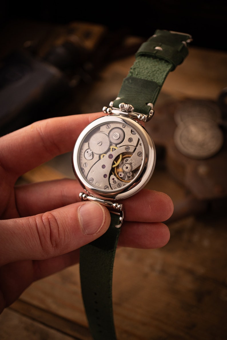 May include: A silver pocket watch with a green leather strap. The watch face is open, revealing the intricate inner workings of the watch.