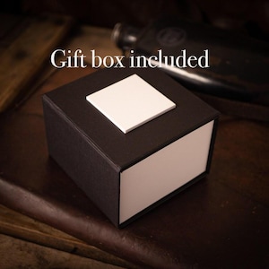 Puede incluir: Una caja de regalo negra con una tapa blanca y un cuadrado blanco en la parte superior. El texto "Gift box included" es visible sobre la caja.