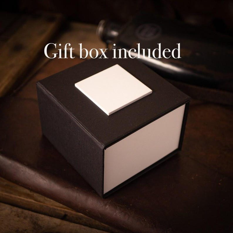 Puede incluir: Una caja de regalo negra con una tapa blanca y un cuadrado blanco en la parte superior. El texto "Gift box included" es visible sobre la caja.