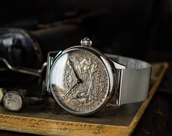 May include: A silver wristwatch with a white face and a silver mesh band. The face of the watch features a detailed engraving of a coat of arms with the text "FIVE SHILLINGS MDCCLXXVII".