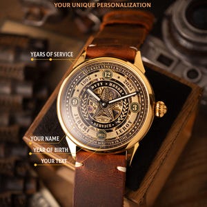 May include: A gold-toned wristwatch with a brown leather strap. The watch face features intricate skeletonized mechanisms, the text "DUTY HONOR", and the words "COURAGE LEAVES NO SCARS". The watch is personalized with the text "JEREMI" and "LEROY".