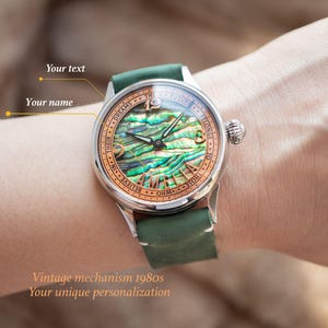 May include: A silver wristwatch with a green leather strap. The watch face has a mother-of-pearl dial with a unique design and the words "IN THEIR DREAMS THE FUTURE BELONGS TO THOSE WHO BELIEVE" around the edge.