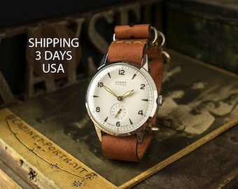 May include: A silver wristwatch with a white face and black hands. The watch has a brown leather band and a small second hand dial. The watch face has the word "СТАРТ" printed on it.