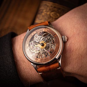 May include: A vintage wristwatch with a skeleton dial, revealing the internal mechanisms. The watch has a brown leather strap and a silver case with copper-toned details. The watch face displays the text "WILLIAM VINTAGE ORIGINAL 1983".