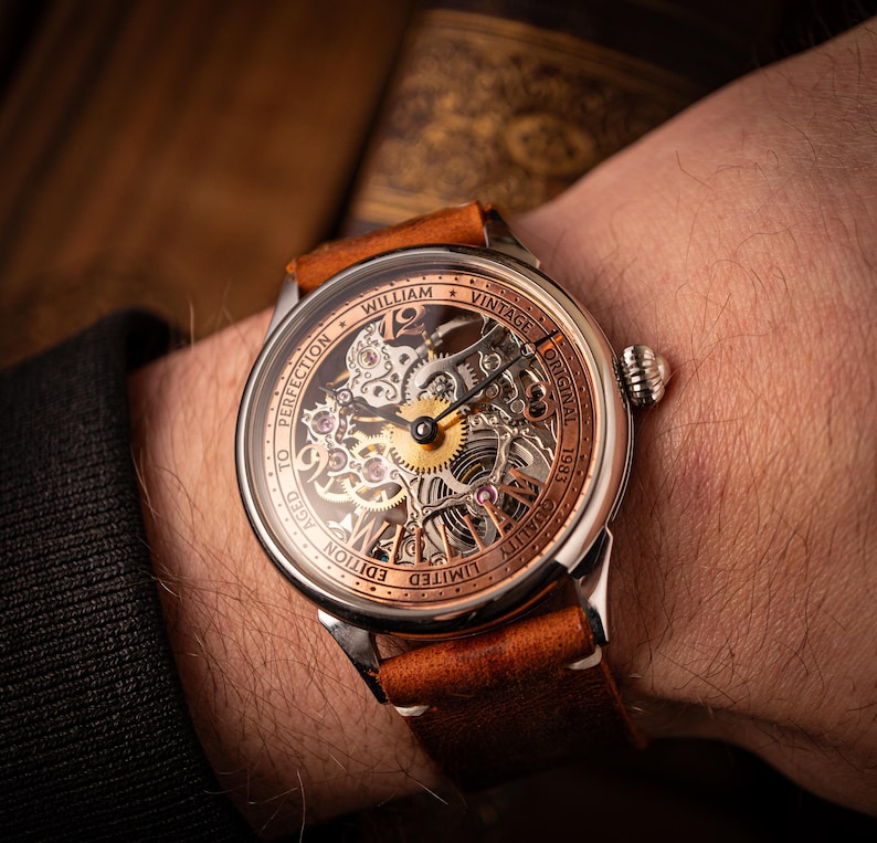 May include: A vintage wristwatch with a skeleton dial, revealing the internal mechanisms. The watch has a brown leather strap and a silver case with copper-toned details. The watch face displays the text "WILLIAM VINTAGE ORIGINAL 1983".