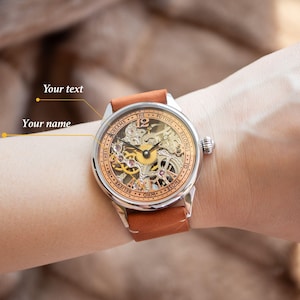 K&ouml;nnte beinhalten: Eine silberne und goldene Skelettuhr mit einem braunen Lederband. Das Zifferblatt der Uhr tr&auml;gt den Text "THE FUTURE BELONGS TO THOSE WHO BELIEVE IN THEIR DREAMS..." am Rand.