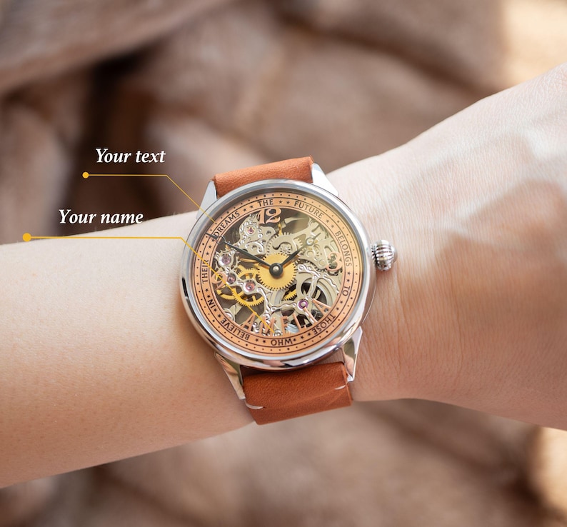 K&ouml;nnte beinhalten: Eine silberne und goldene Skelettuhr mit einem braunen Lederband. Das Zifferblatt der Uhr tr&auml;gt den Text "THE FUTURE BELONGS TO THOSE WHO BELIEVE IN THEIR DREAMS..." am Rand.