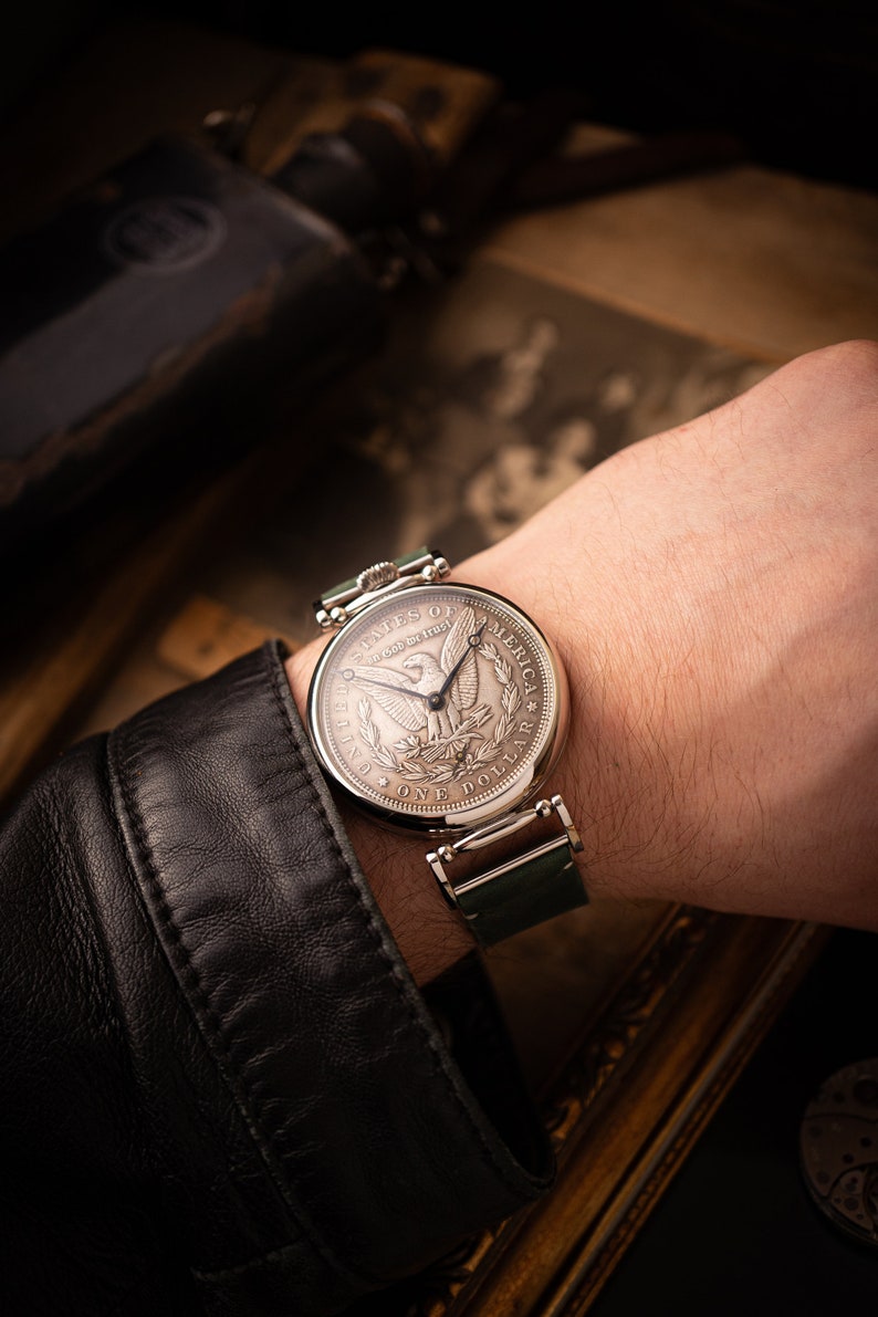 May include: A silver watch with a green leather band. The face of the watch is a replica of a United States silver dollar coin with the words "United States of America" and "One Dollar" inscribed on the face.