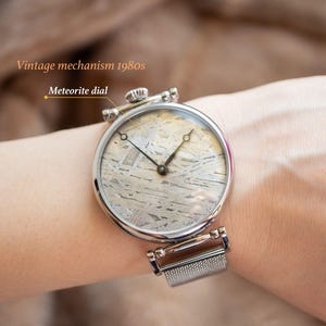 May include: A silver wristwatch with a mesh band and a round, silver face. The watch face has a unique meteorite dial with a black hour and minute hand. The text "Vintage mechanism 1980s" and "Meteorite dial" are visible in the image.