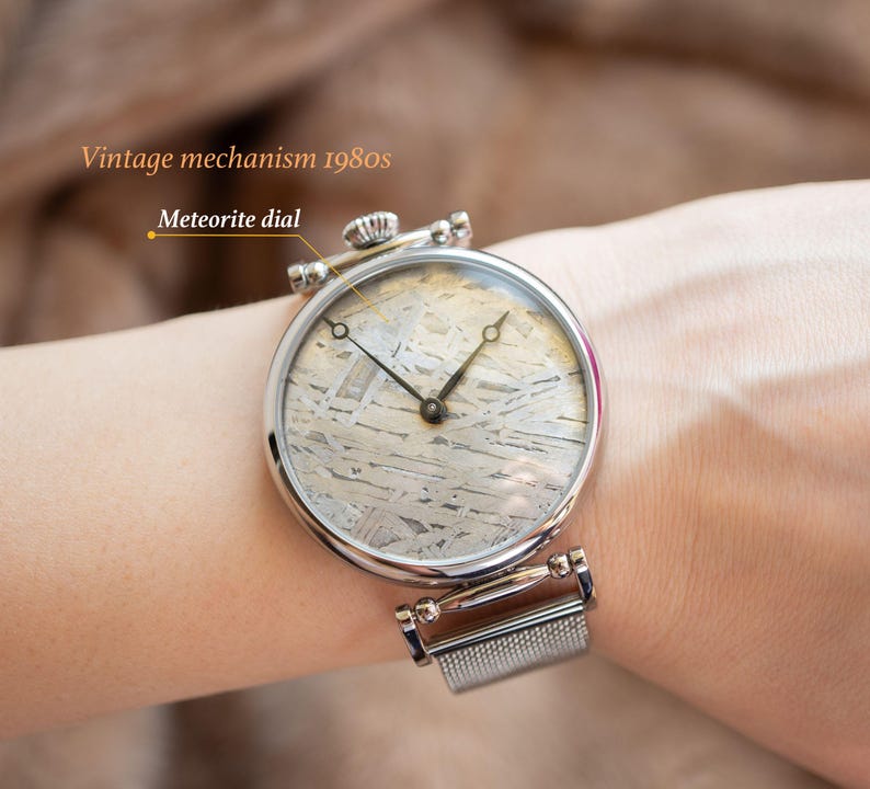 May include: A silver wristwatch with a mesh band and a round, silver face. The watch face has a unique meteorite dial with a black hour and minute hand. The text "Vintage mechanism 1980s" and "Meteorite dial" are visible in the image.