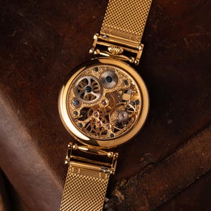 May include: A gold-toned skeleton watch with a mesh band. The watch face reveals intricate gears and jewels. The watch is set against a dark brown leather surface, highlighting its elegant design.