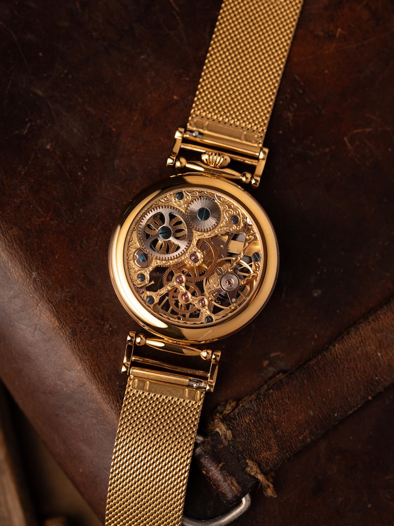 May include: A gold-toned skeleton watch with a mesh band. The watch face reveals intricate gears and jewels. The watch is set against a dark brown leather surface, highlighting its elegant design.