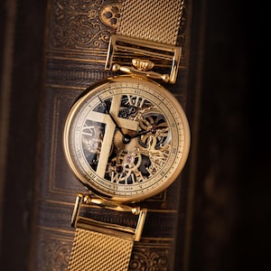May include: A gold-toned wristwatch with a mesh band, resting on a textured, dark brown surface. The watch face features Roman numerals and visible internal gears. The text "VII Garrison VI 1919" is visible on the watch face.