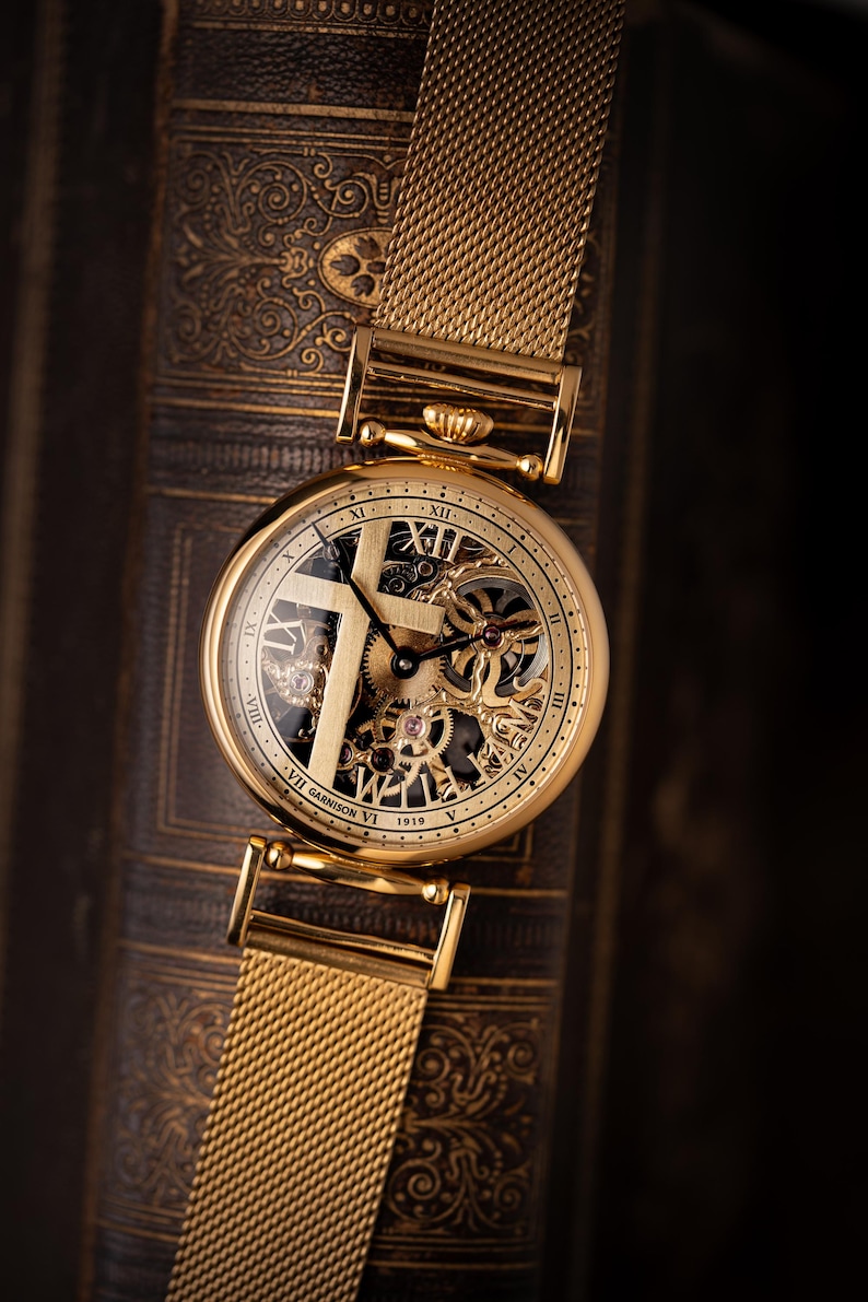 May include: A gold-toned wristwatch with a mesh band, resting on a textured, dark brown surface. The watch face features Roman numerals and visible internal gears. The text "VII Garrison VI 1919" is visible on the watch face.