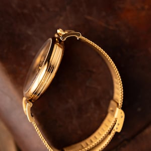 May include: A gold-toned wristwatch with a mesh band. The watch face is round and the band is a fine, woven mesh. The watch is set against a dark, textured background, highlighting its metallic sheen. The watch band is approximately 7 inches in length.