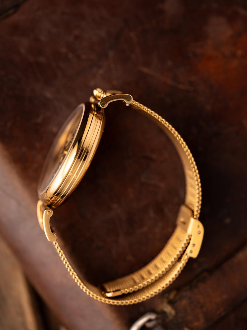 May include: A gold-toned wristwatch with a mesh band. The watch face is round and the band is a fine, woven mesh. The watch is set against a dark, textured background, highlighting its metallic sheen. The watch band is approximately 7 inches in length.