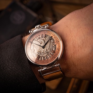 May include: A silver wristwatch with a round, copper-colored face. The face features a silver eagle with outstretched wings and the text "The Future Belongs To Those Who Believe In Their Dreams 1983".