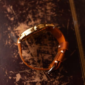 May include: A gold wristwatch with a brown leather strap. The watch's side profile shows a textured crown. The strap has white stitching and a silver buckle. The watch is set against a dark, textured background. The strap is approximately 2 cm wide.