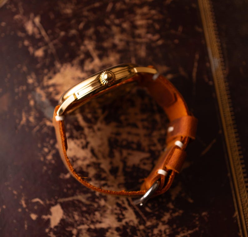 May include: A gold wristwatch with a brown leather strap. The watch's side profile shows a textured crown. The strap has white stitching and a silver buckle. The watch is set against a dark, textured background. The strap is approximately 2 cm wide.
