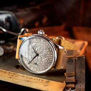 Puede incluir: Un reloj de bolsillo plateado con una correa de cuero marrón. La esfera del reloj presenta un diseño detallado de una figura histórica y el texto "E PLURIBUS UNUM 1885".
