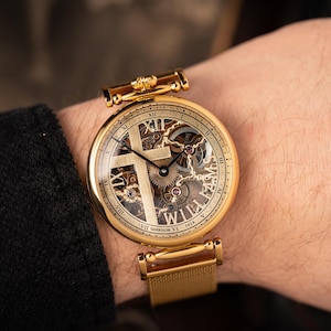 May include: Gold-toned wristwatch with a skeleton dial featuring a cross design. The watch has Roman numeral hour markers and visible gears. The band is a gold mesh style. The watch face includes the text "WILLIAMS" and "GARRISON VI 1919".