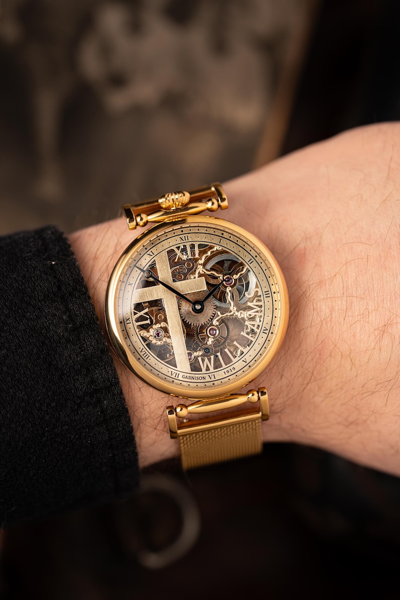 May include: Gold-toned wristwatch with a skeleton dial featuring a cross design. The watch has Roman numeral hour markers and visible gears. The band is a gold mesh style. The watch face includes the text "WILLIAMS" and "GARRISON VI 1919".