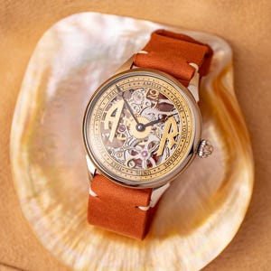 K&ouml;nnte beinhalten: Eine goldfarbene Armbanduhr mit einem skelettierten Zifferblatt und einem braunen Lederarmband. Das Zifferblatt tr&auml;gt die Aufschrift "Chase Your Dreams Until They Become Your Reality" und "Amelia".