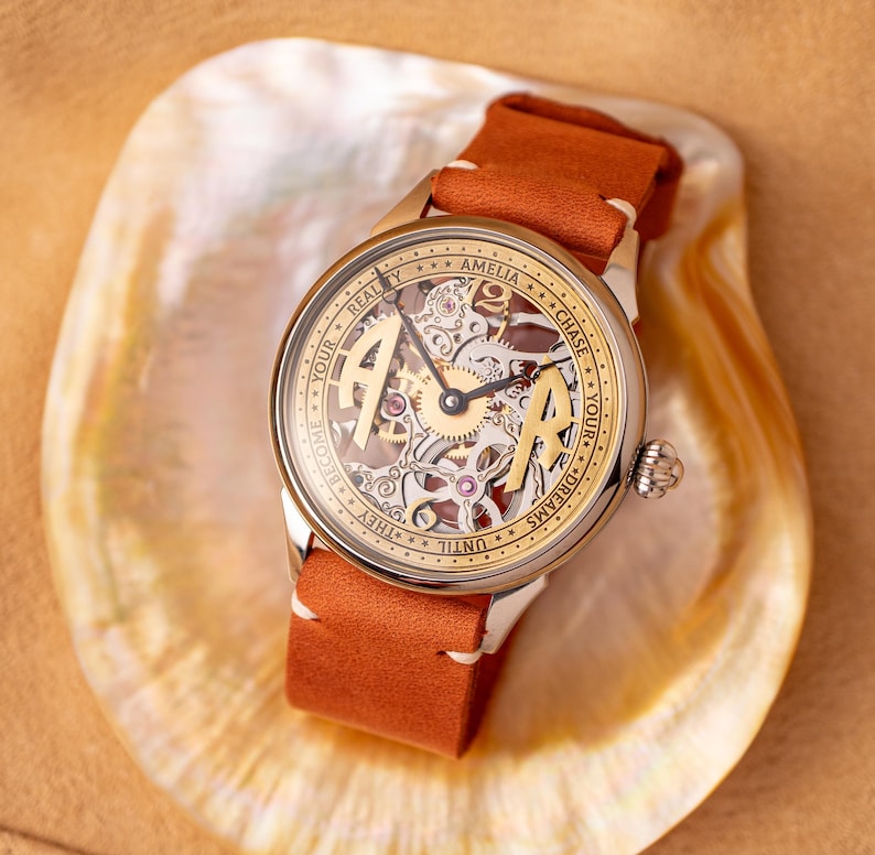 K&ouml;nnte beinhalten: Eine goldfarbene Armbanduhr mit einem skelettierten Zifferblatt und einem braunen Lederarmband. Das Zifferblatt tr&auml;gt die Aufschrift "Chase Your Dreams Until They Become Your Reality" und "Amelia".