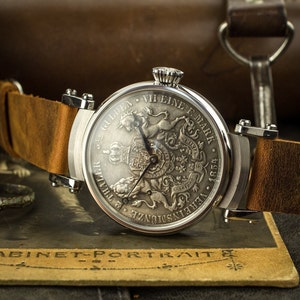 Puede incluir: Un reloj de bolsillo de plata con una correa de cuero marrón. La esfera del reloj presenta un grabado detallado de un escudo de armas con un unicornio y el texto "5. GOLDEN VI. EINE F. MARK 1891."