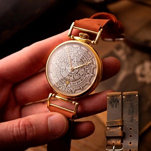 Puede incluir: Un reloj de bolsillo dorado con una cara blanca circular detallada que presenta números romanos y un diseño similar a un calendario. El reloj tiene una correa de cuero marrón y se sostiene en una mano.